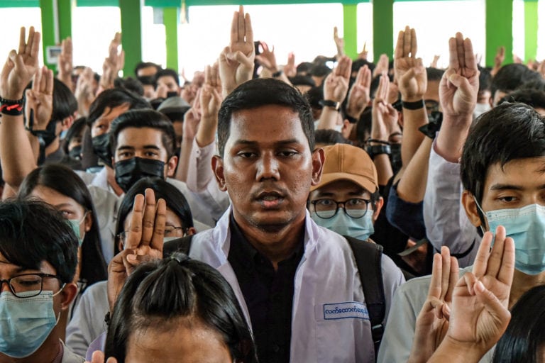 A group of inidividuals are pictures in lab coasts. Most are holding up there hands with three fingers up, signifying the civil disobedience movement in Myanmar